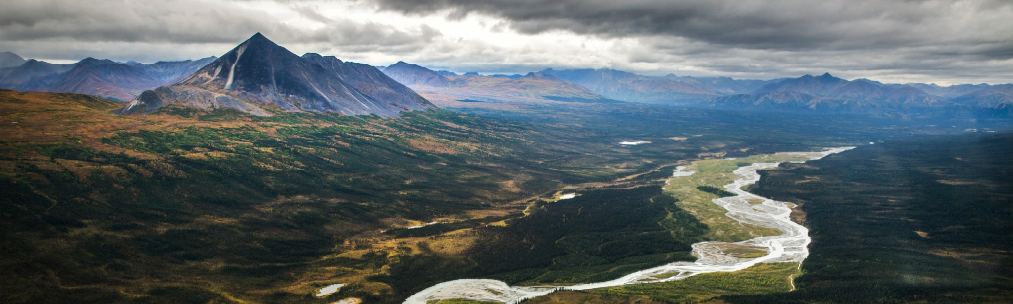 expansive aerial view of a twisting and winding river stretching through a forested valley beneath dark clouds with a mountain peak on the left