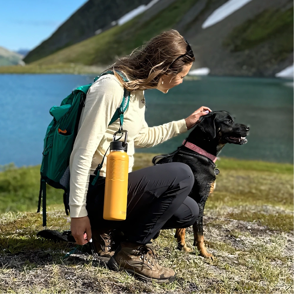 Female hiker kneeling beside black dog in front of alpine lake, with goldenrod Nuataaq Carabiner Bottle clipped to green backpack