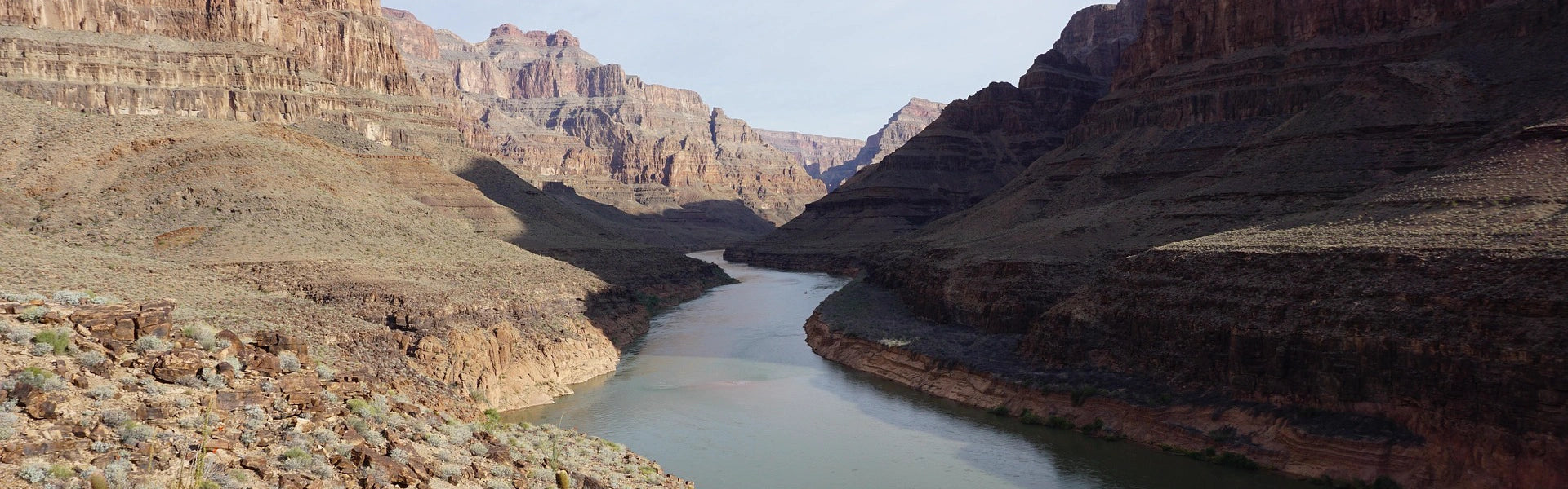 wide dusty gray river cutting through a weather worn mountain gorge with small dessert brush covering the sides of the gorge