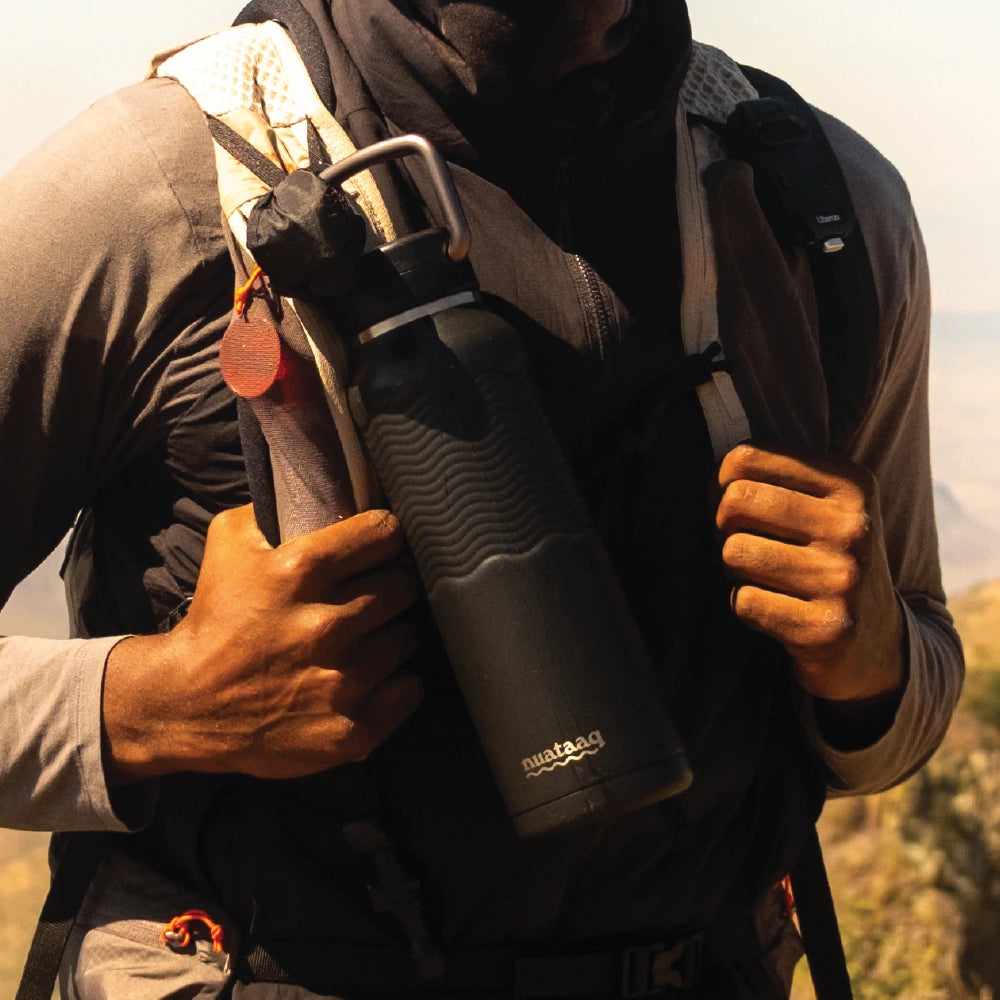 Person holding a black nuataaq carabiner bottle water bottle with a backpack outdoors