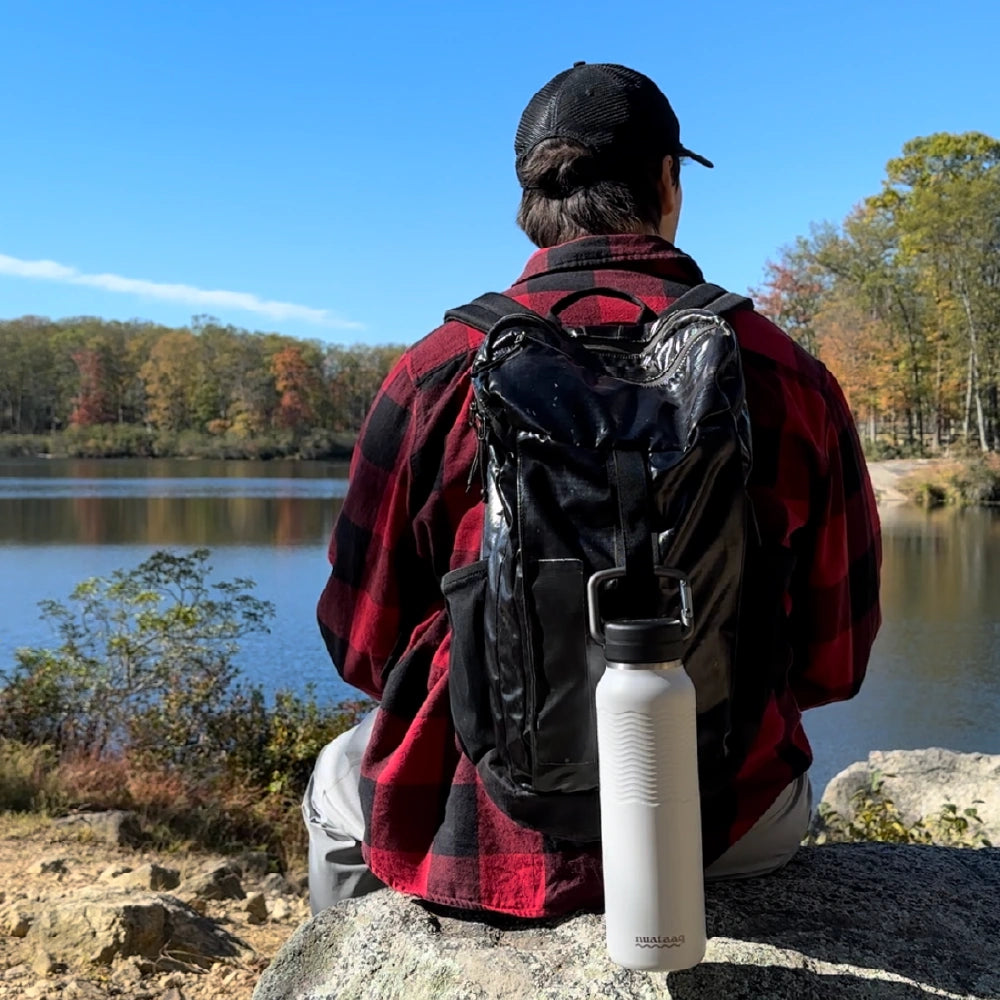 Mist gray Nuataaq connect™ carabiner bottle attached to a mans backpack as he sits on a large rock overlooking a forested lake
